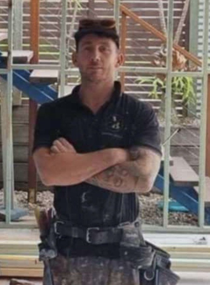 A man with crossed arms standing outdoors, wearing a black polo shirt, with construction or play equipment and wooden structures in the background.