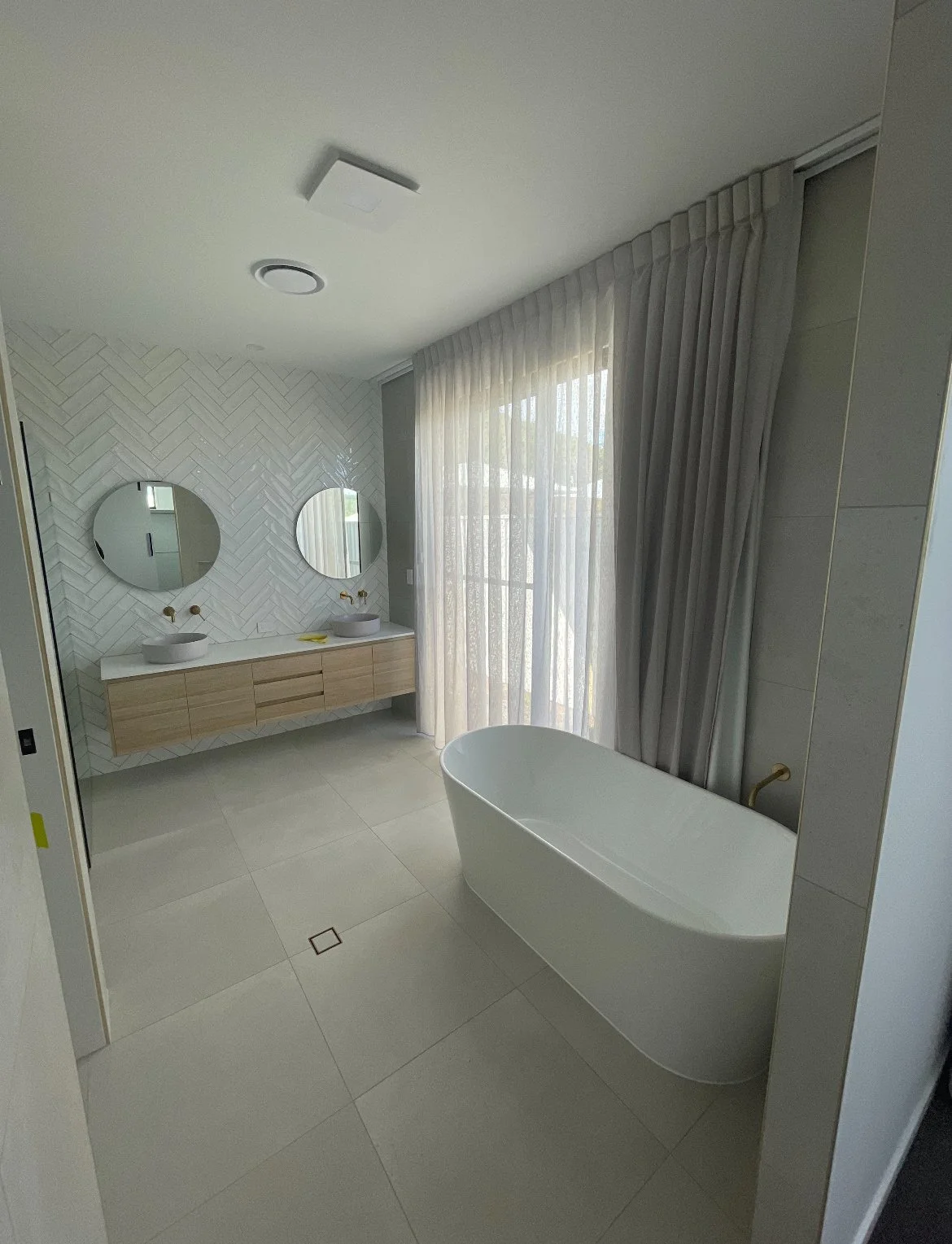 Modern bathroom with a white bathtub, a floating wooden vanity with two vessel sinks, two round mirrors, and large window with sheer curtains.