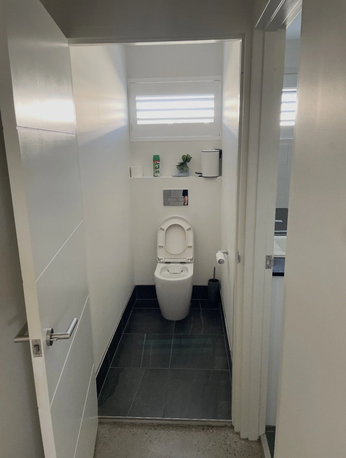 Small modern bathroom with a toilet, toilet paper, a toilet brush, a window, and some bathroom accessories on a shelf.