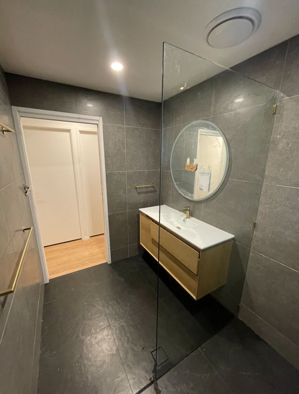 Modern bathroom with gray tiled walls and floor, wooden vanity with sink, round mirror, and a glass dividing wall.