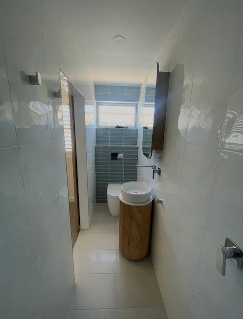 A modern bathroom with a narrow layout featuring a white sink, a wooden cabinet, a mirror, a toilet, a small window, and white tiled walls and floors.