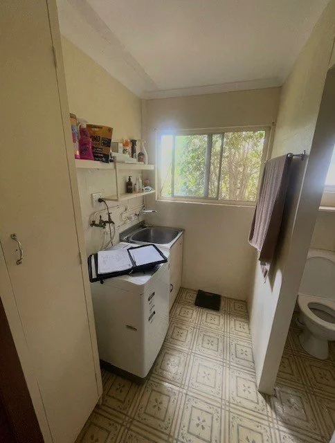 Small laundry room with a washing machine and a sink, open shelves with cleaning supplies and other items, a window showing trees outside, and a toilet partially visible.