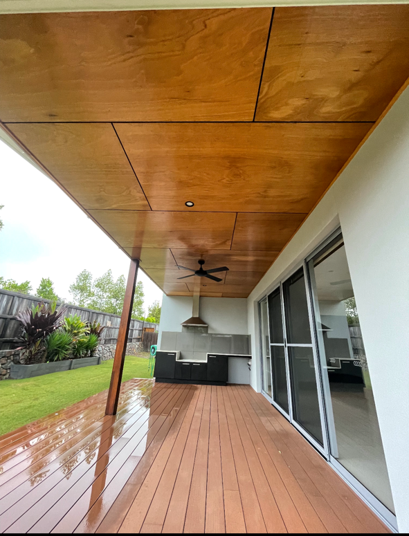 Covered outdoor patio with stained wood ceiling, ceiling fan, and built-in grill area, adjacent to a glass sliding door, in backyard with green grass and plants.