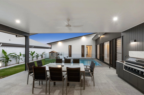Covered outdoor patio with dining table, chairs, a pool, and barbecue grill, adjacent to modern house with glass doors and windows, outdoor lighting, and a backyard with palm trees.