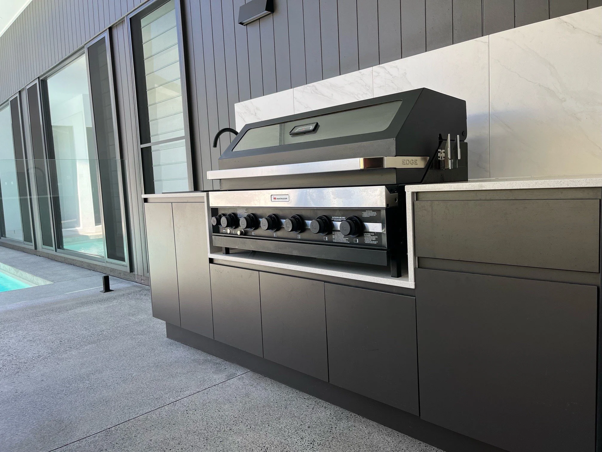 Outdoor modern stainless steel grill with a closed lid, situated on a concrete patio next to a building with large glass windows.