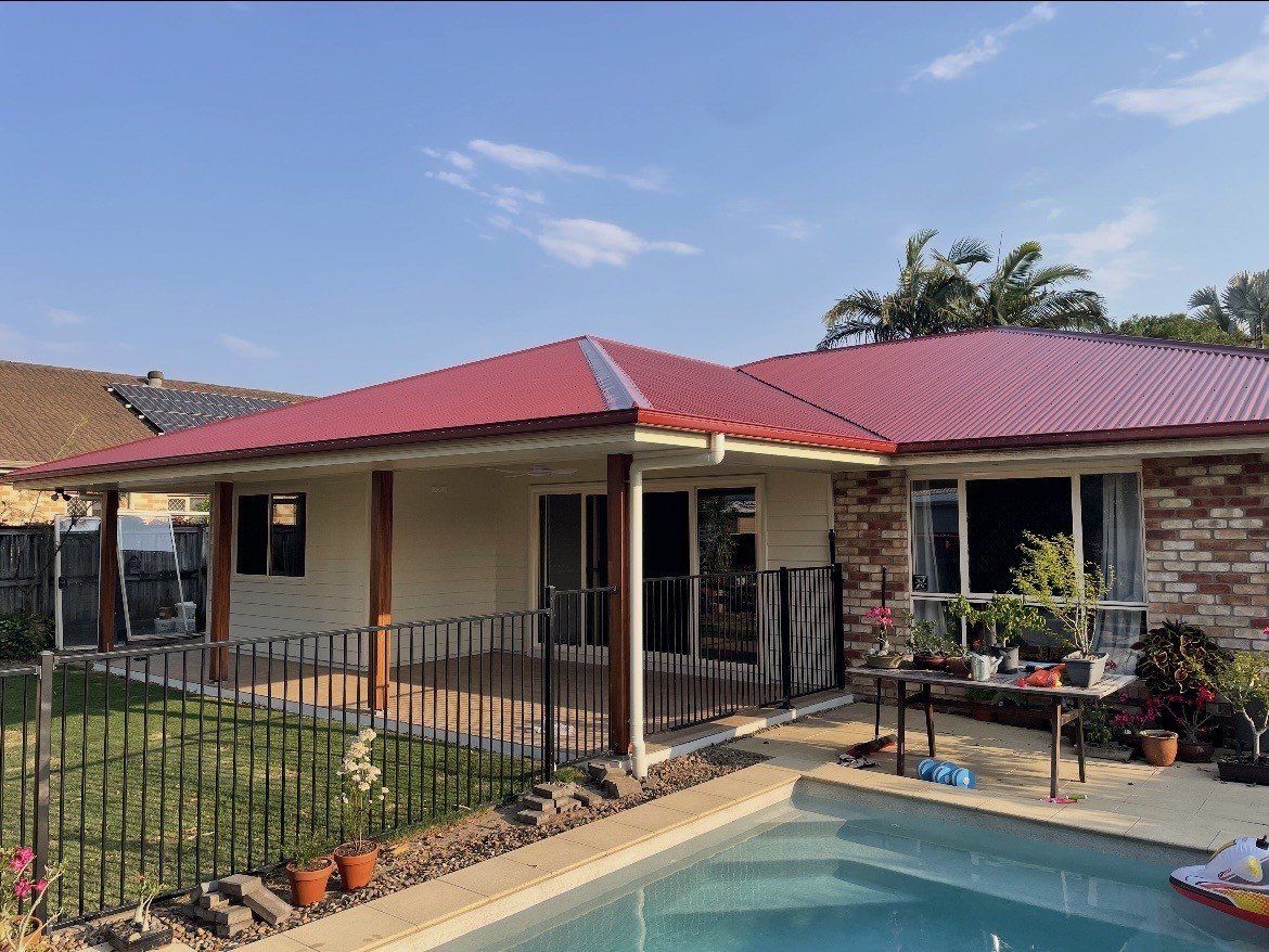 Backyard view of a house with a red metal roof, a small pool, and a fenced patio area with potted plants and gardening supplies.