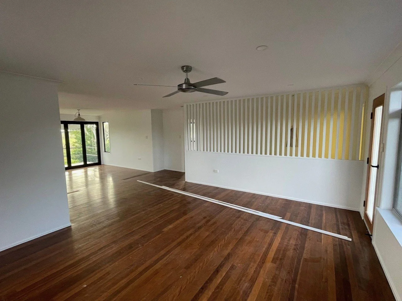 Empty living room with hardwood floors, ceiling fan, and sliding glass door leading outside, with yellow and white striped accent wall.