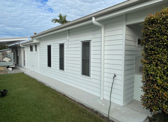 Side view of a white residential house with three closed windows, black security screens, a white gutter downspout, and a green shrub on the right side.