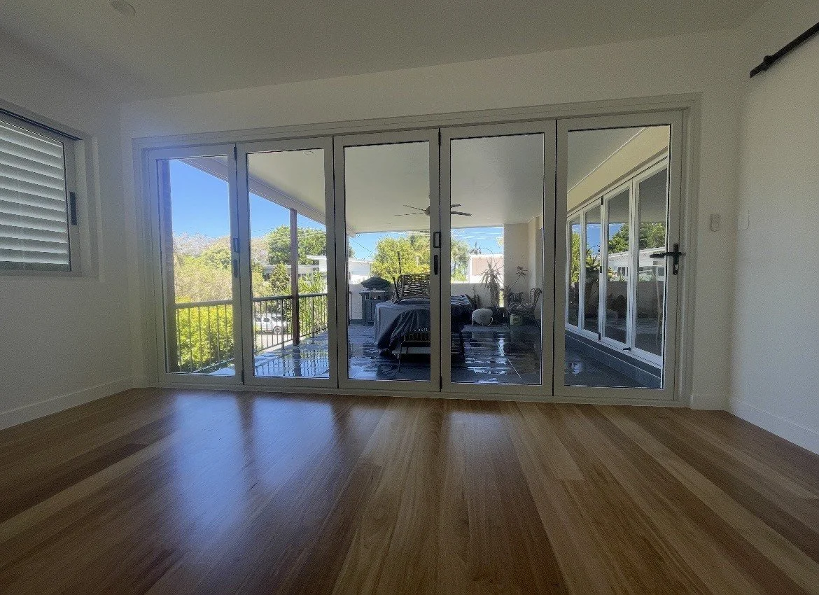 Interior view of a room with a wooden floor and large glass sliding doors leading to a balcony with trees and blue sky visible outside.