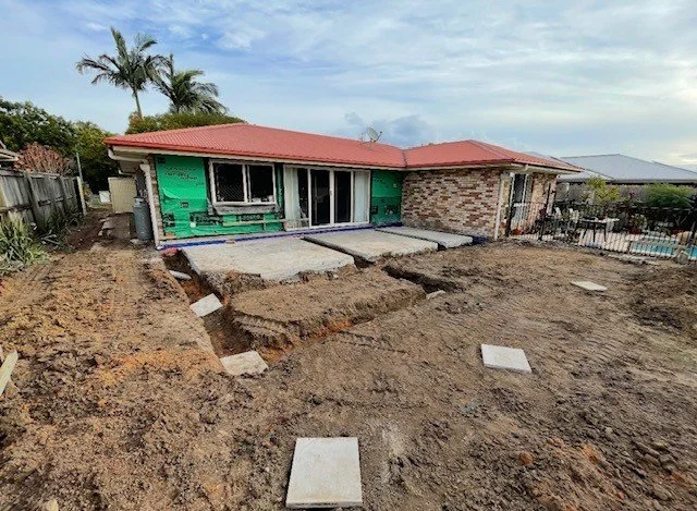 Backyard of a house under construction, with exposed dirt, a concrete patio, and a partially finished exterior wall.