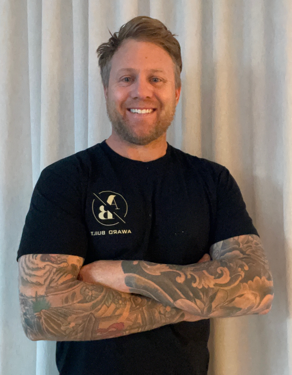A smiling man with tattoos on both arms, wearing a black T-shirt with a logo and text, standing with arms crossed in front of light-colored curtains.
