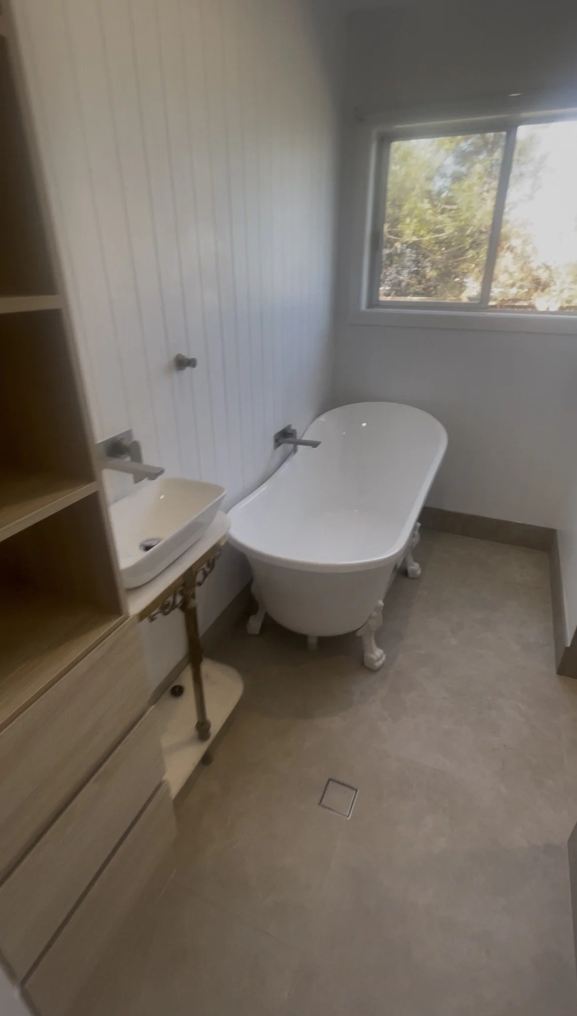 A bathroom with a clawfoot bathtub near a window, a small vessel sink on a wooden vanity, and built-in shelves on the left side.