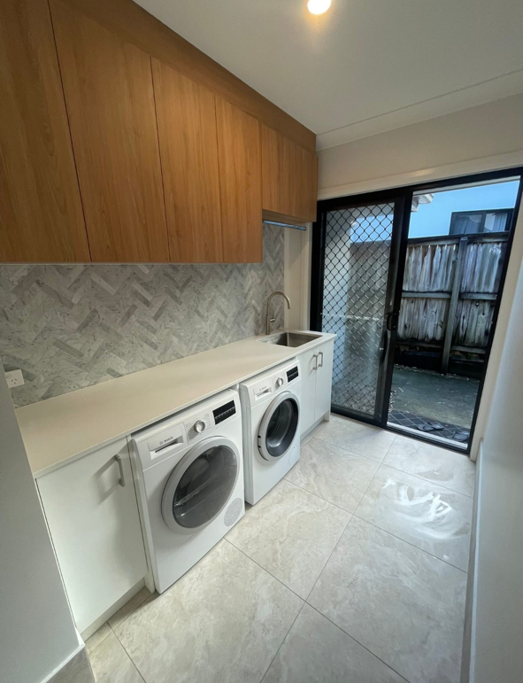 Laundry room with white front-loading washing machine and dryer, wooden overhead cabinets, a gray patterned backsplash, a sink, and a sliding glass door leading outside.