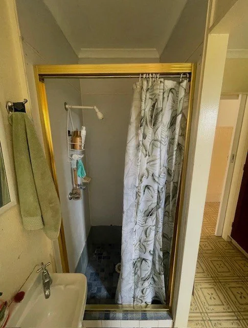 Small enclosed shower with a patterned curtain, a wall-mounted soap dish, and a showerhead, adjacent to a bathroom sink with a green towel hanging on a nearby towel bar.