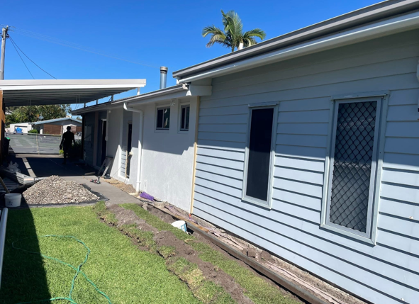 Side view of a house with white siding, three windows, and a small yard with grass and a garden bed under a clear blue sky.
