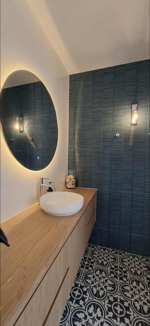 Modern bathroom with a round mirror illuminated by backlighting, a white vessel sink on a wooden vanity, black textured wall tiles, patterned floor tiles, and two wall-mounted light fixtures.