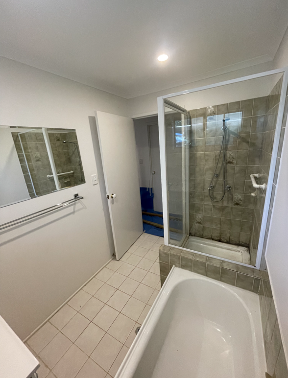 A small bathroom with white walls, beige tiled floor, a bathtub in the foreground, a shower area with a glass door, mirror, and towel rack on the wall, and an open door leading to a hallway or storage area.
