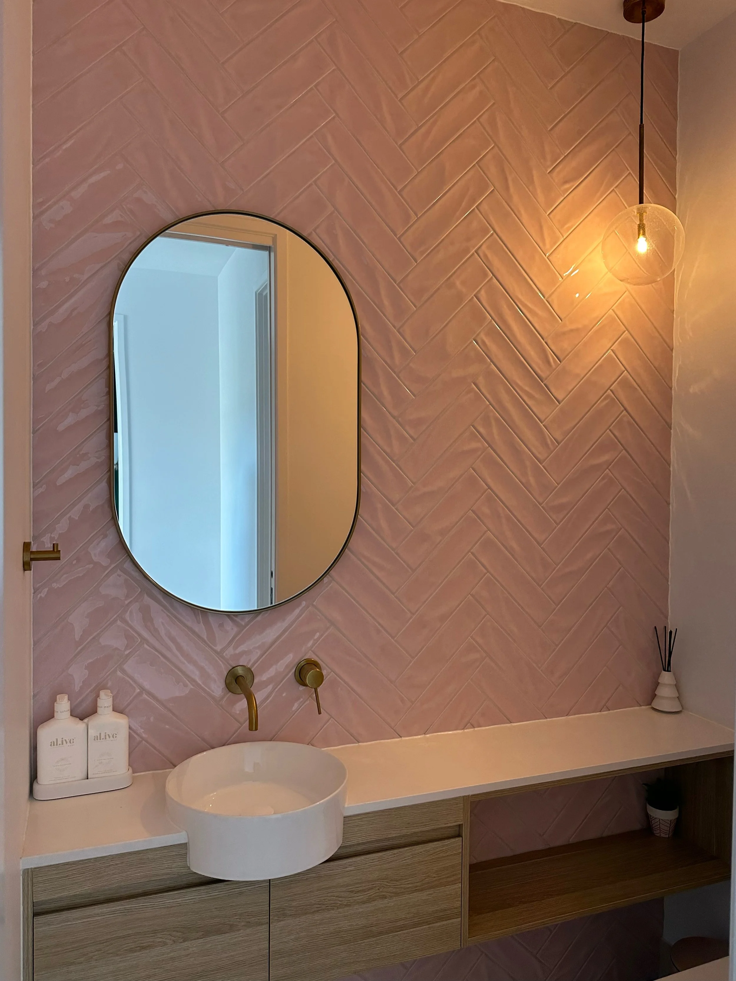 Modern bathroom vanity with a round vessel sink, wall-mounted faucet, and a large oval mirror against a pink herringbone tile wall, illuminated by a pendant light, with decorative bottles and a reed diffuser on the counter.