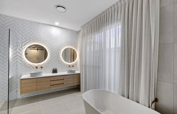 Modern bathroom with two round illuminated mirrors, vessel sinks, wooden vanity, white textured wall, large window with sheer curtains, and a freestanding bathtub.