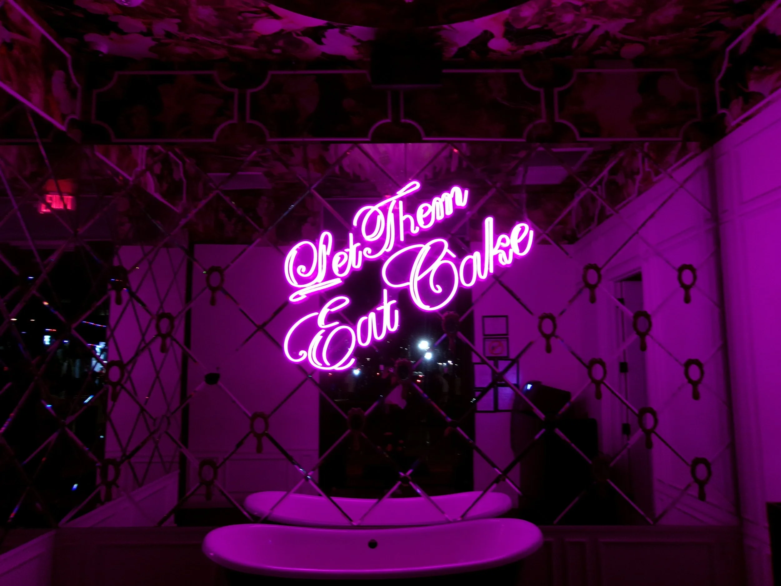 Pink neon sign reading 'Set Them Eat Cake' in front of a decorative mirror wall with a reflection of a person. The room is illuminated with pink and purple lighting.