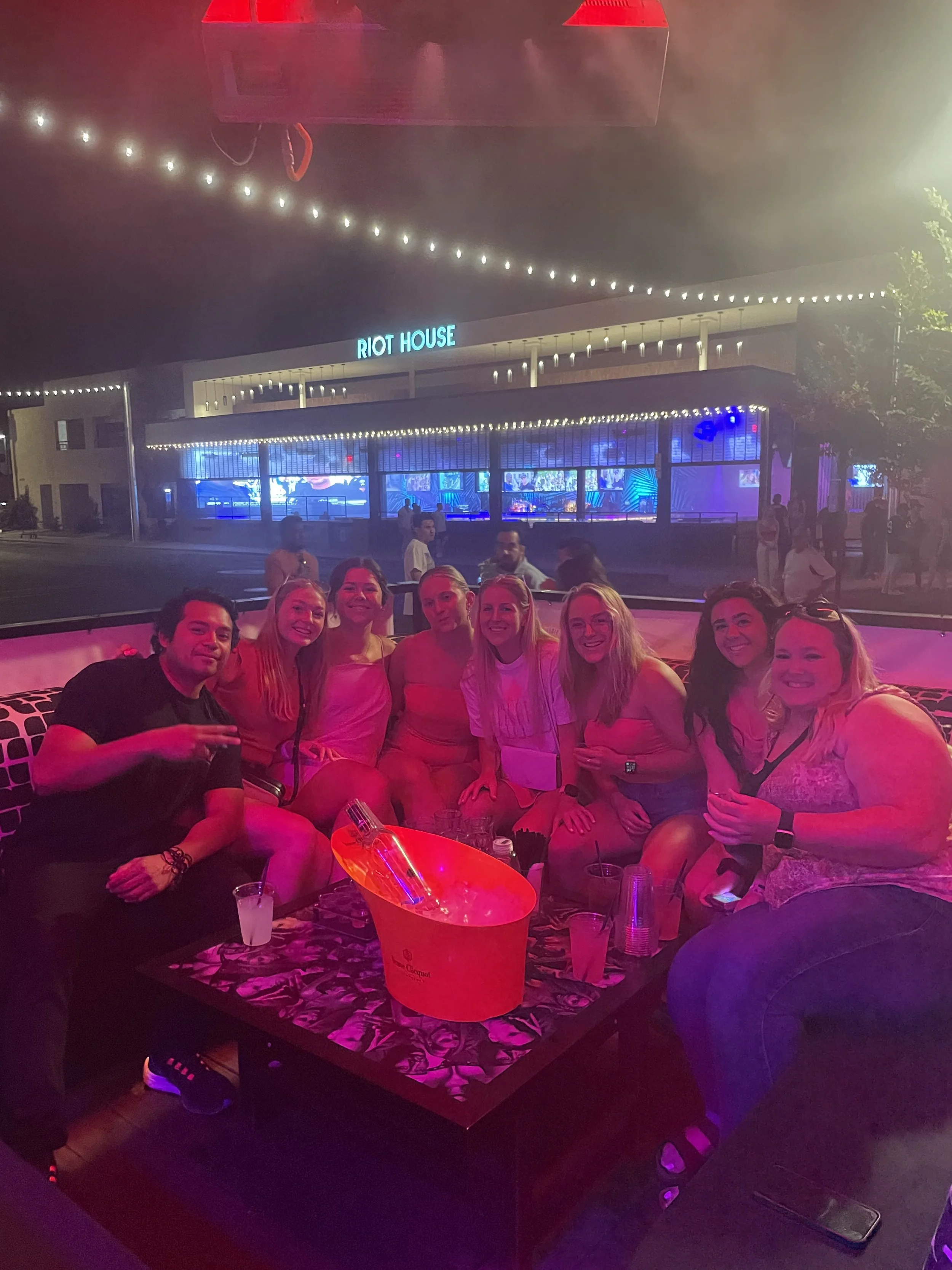 A group of nine people sitting together at a table outside of Riot House nightclub, illuminated by pink and purple lights, with a neon sign and purple lighting inside, at night.