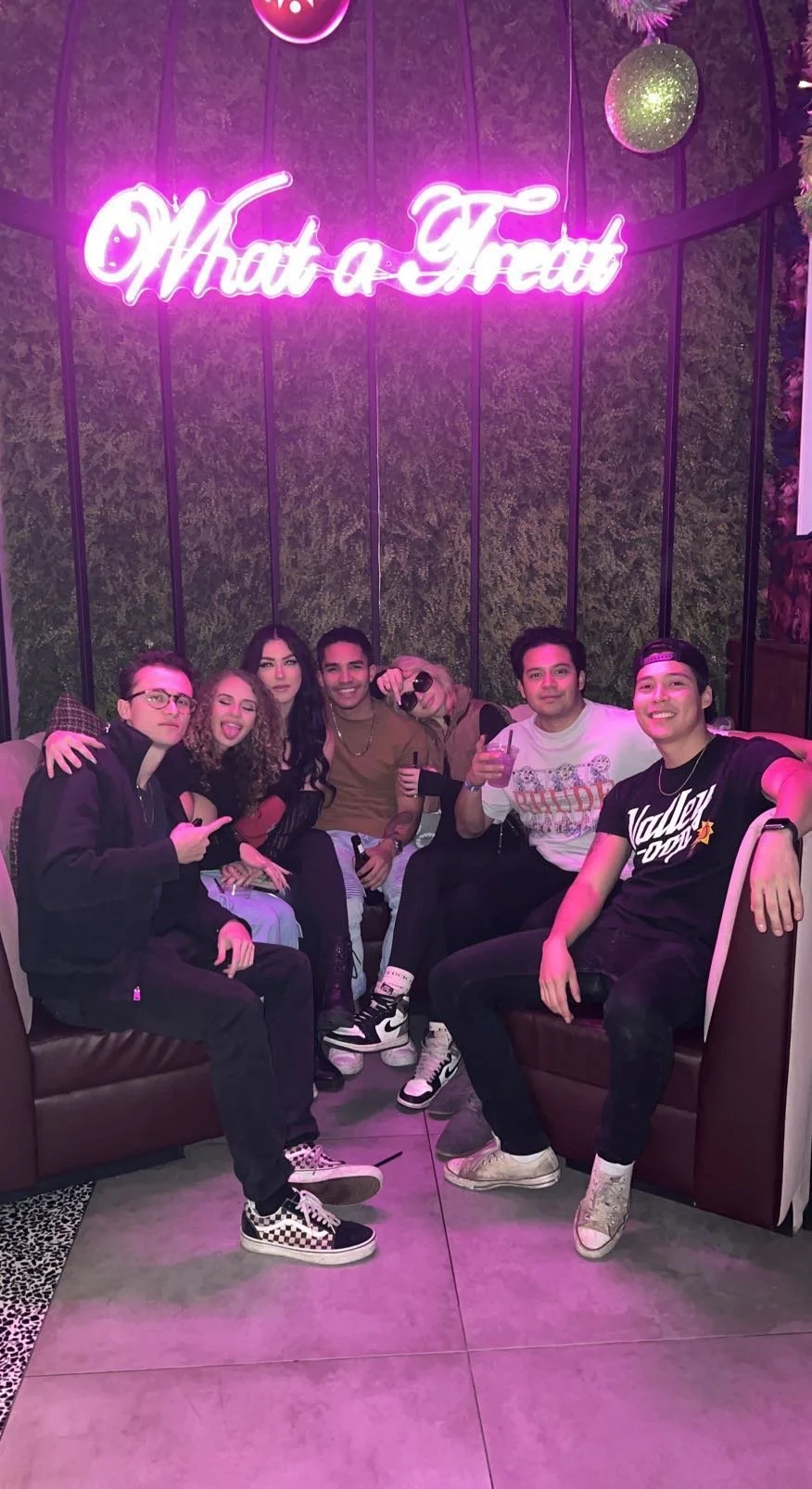 A group of seven young people sitting on sofas against a decorative wall with a glowing pink neon sign that says 'What a Treat'. They are smiling, holding drinks, and posing for the photo.