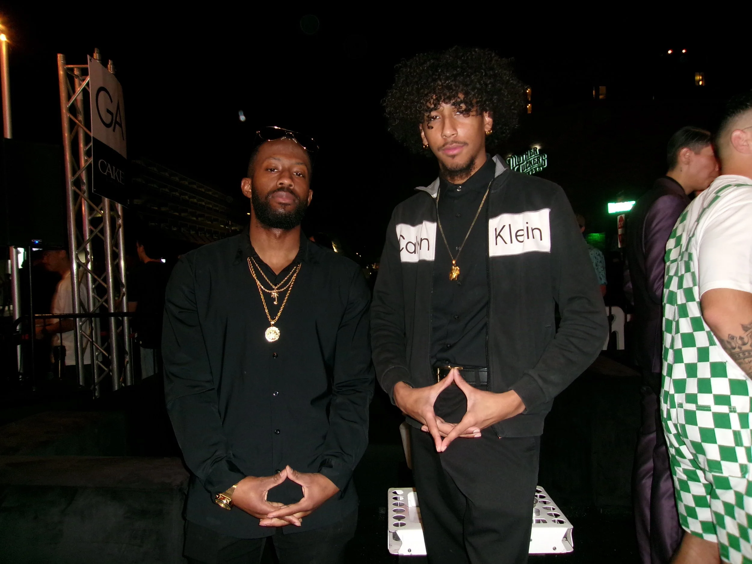 Two men standing together at an indoor event, dressed in black clothing with gold jewelry, with a DJ booth and other people in the background.