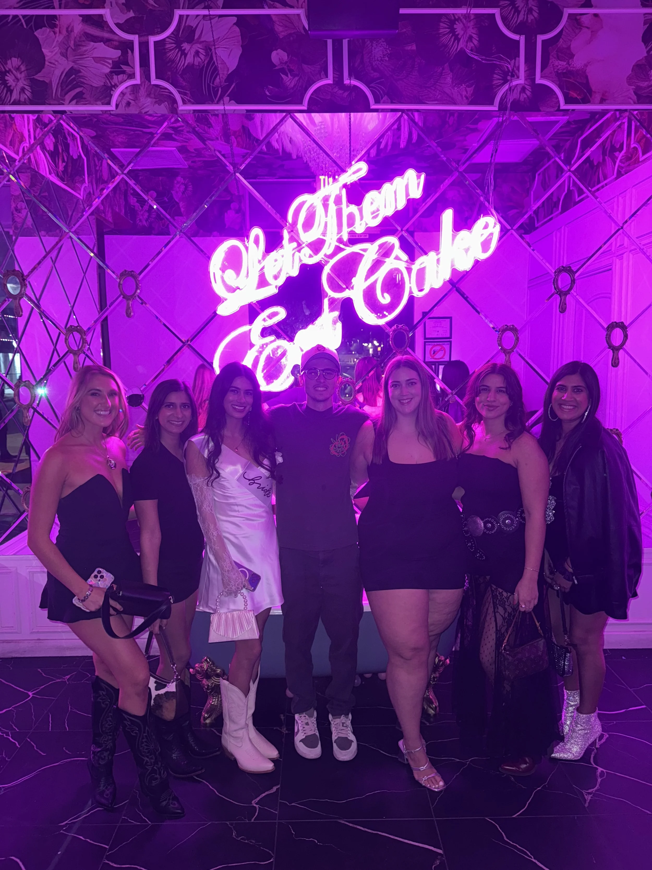 Group of seven people posing in front of bright pink neon sign that reads 'Let's Have Some Fun' at a lively party or nightclub.