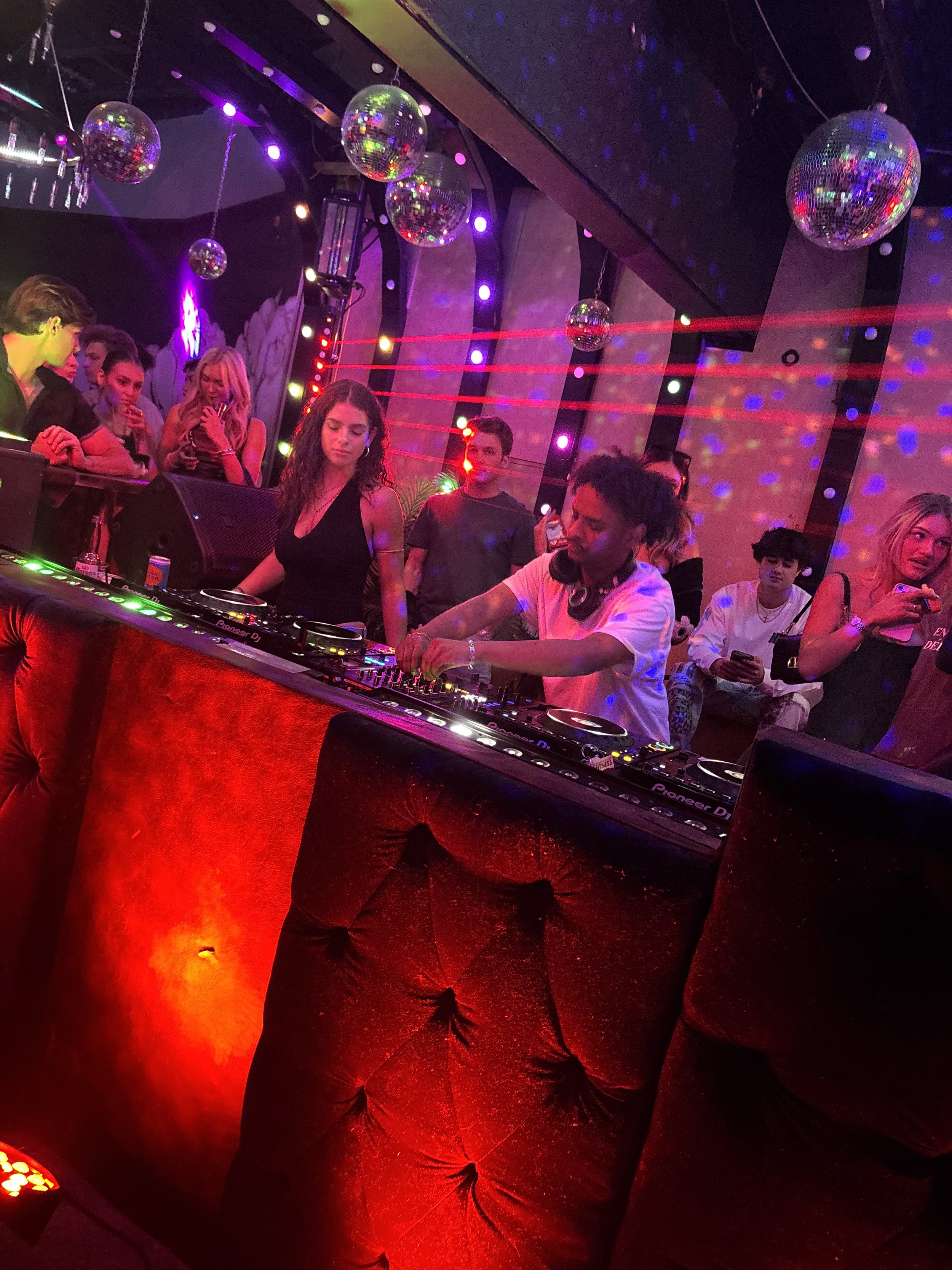 A DJ is performing at a nightclub with a crowd dancing and socializing, colorful lights, and disco balls overhead.