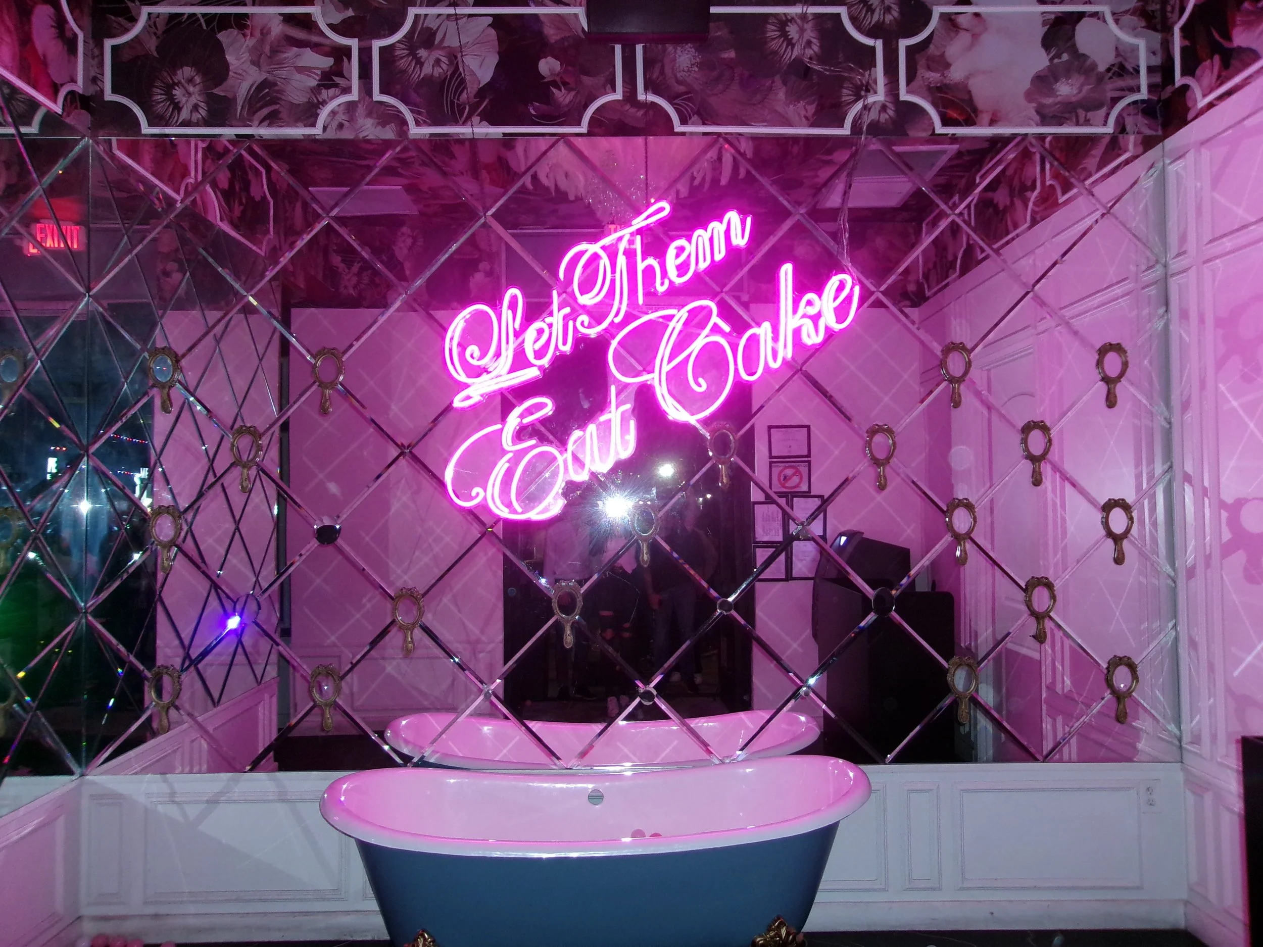 A bright pink neon sign reads 'Let Them Eat Cake' in a decorative script, mounted on a wall with a mirrored lattice pattern, with a bathtub in the foreground and reflection of people and lights in the background.