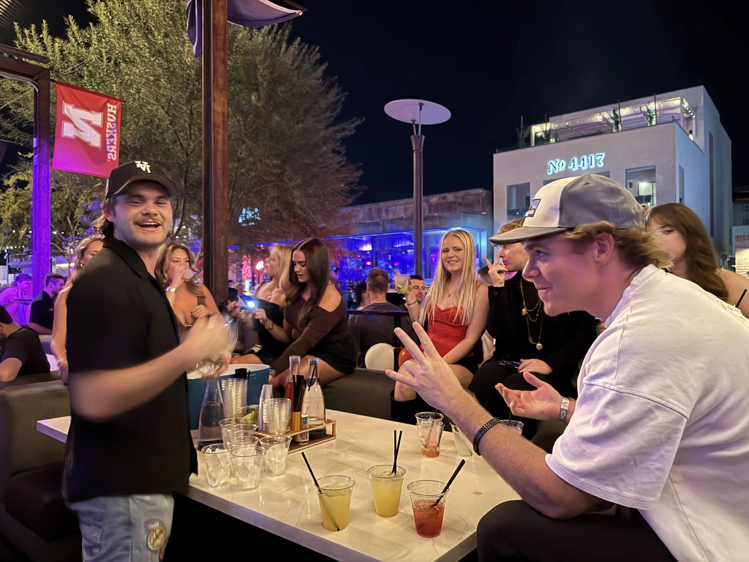 A group of young people socializing at an outdoor bar or club during nighttime. They are sitting around a table with drinks, and one person is standing and related to the camera. The atmosphere has colorful lighting, and in the background, there are trees, a building with neon lights, and other people.