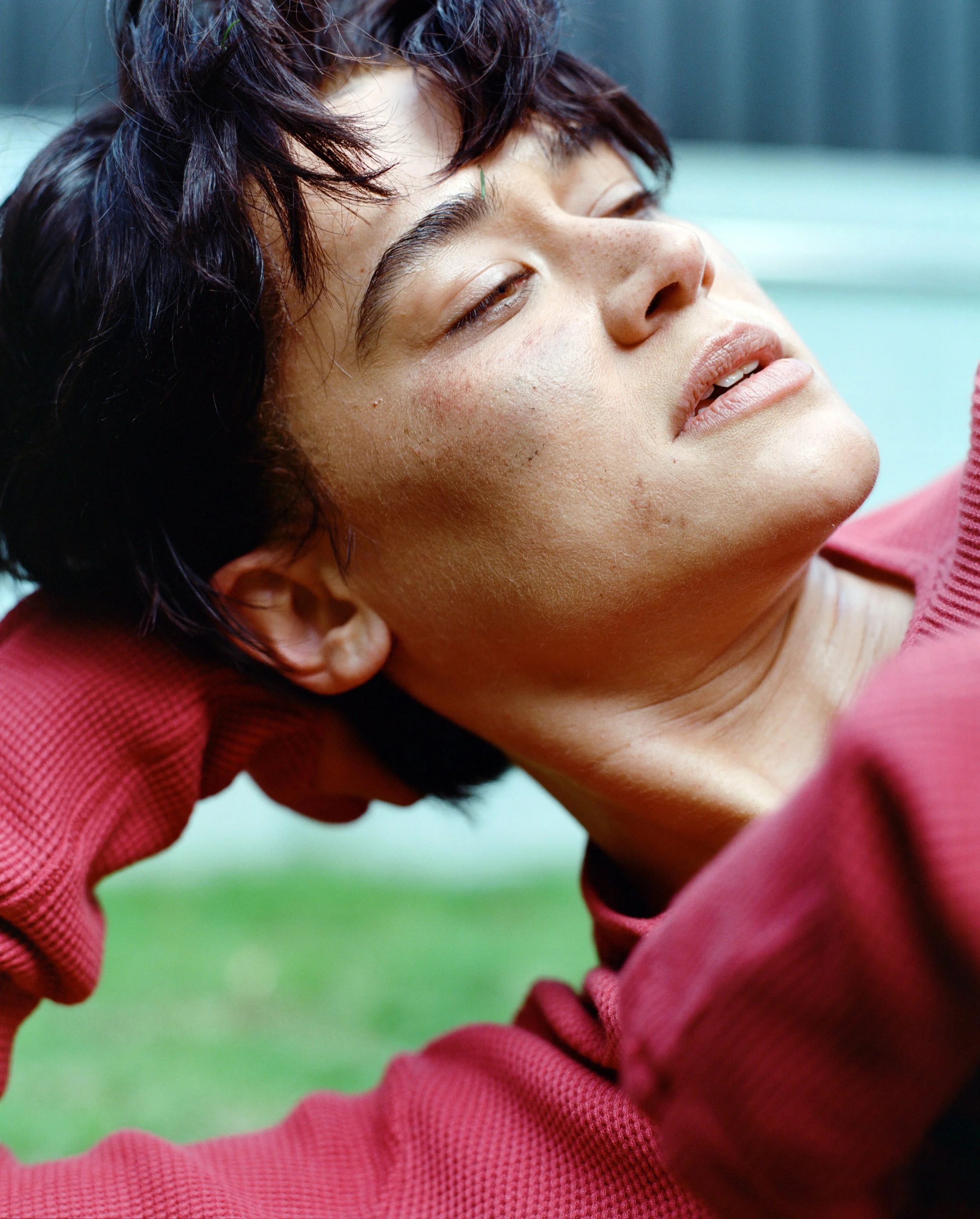 Close-up of Katy O'Brian, wearing a red top, lying outdoor with her eyes partially closed and a relaxed expression.