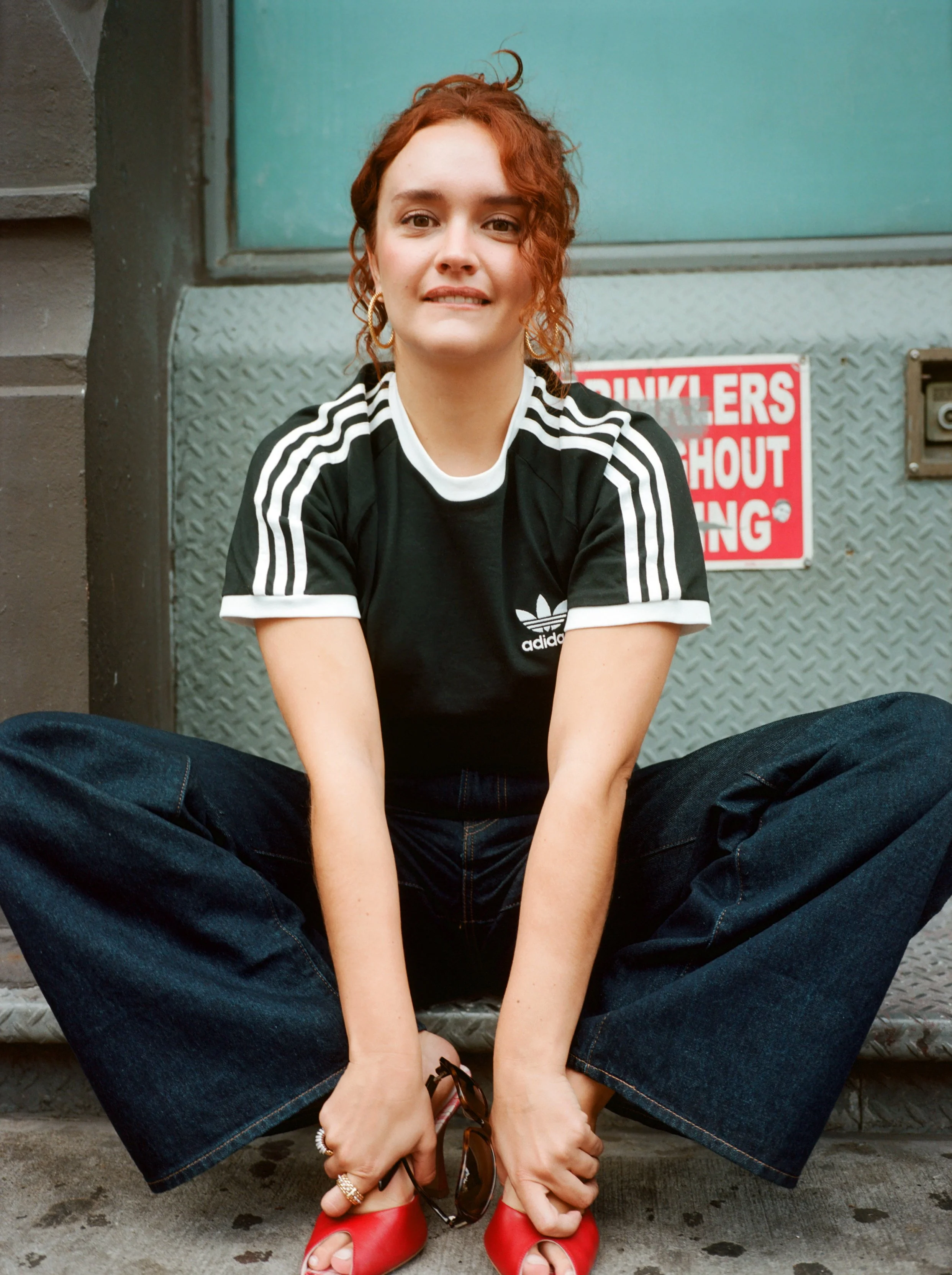 Olivia Cooke sitting on the steps of a building. She's wearing a black Adidas t-shirt, dark jeans, and red open-toe heels, holding glasses in her hands.