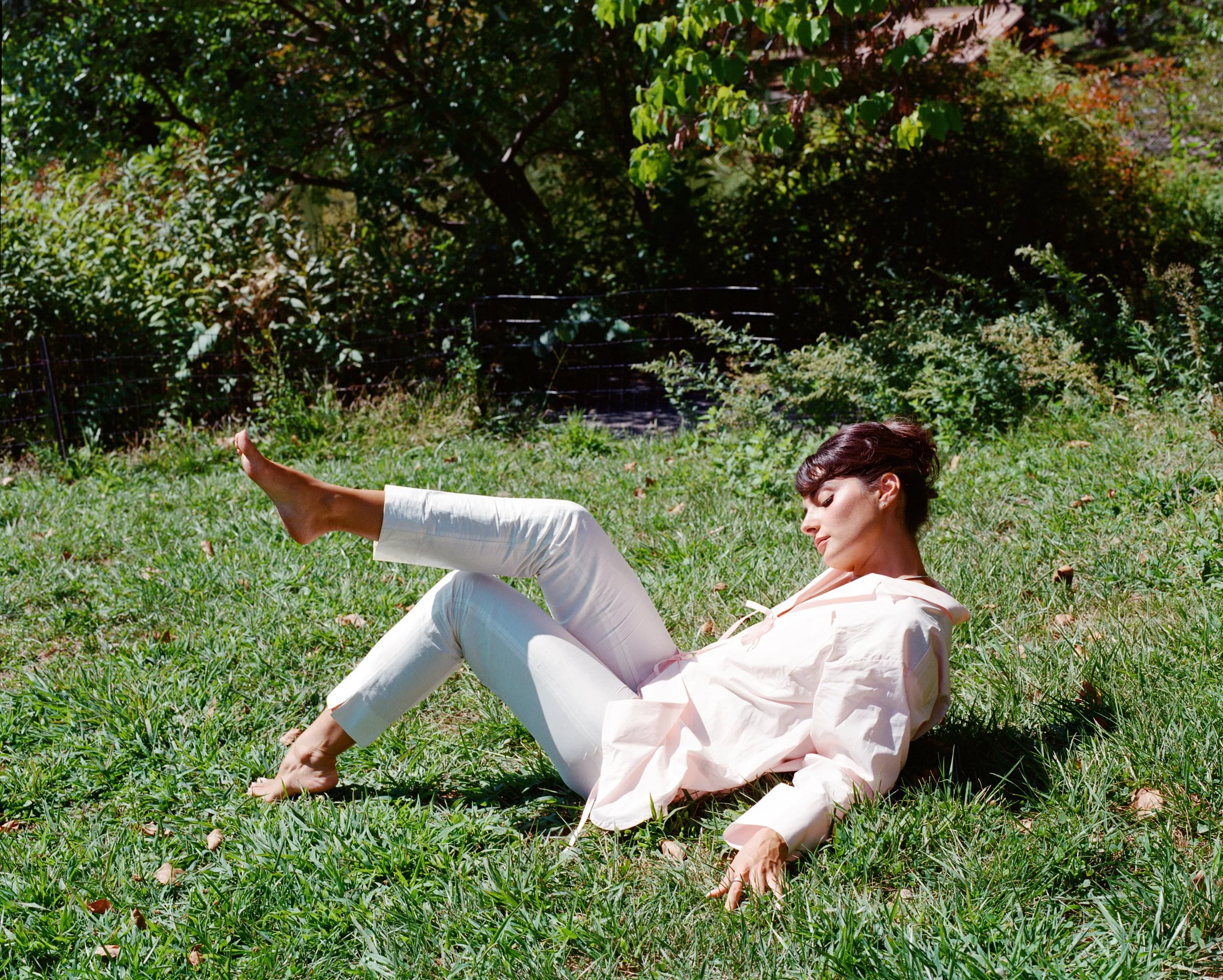 Woman lying on green grass in a garden, wearing a light pink shirt and white pants, with her eyes closed.