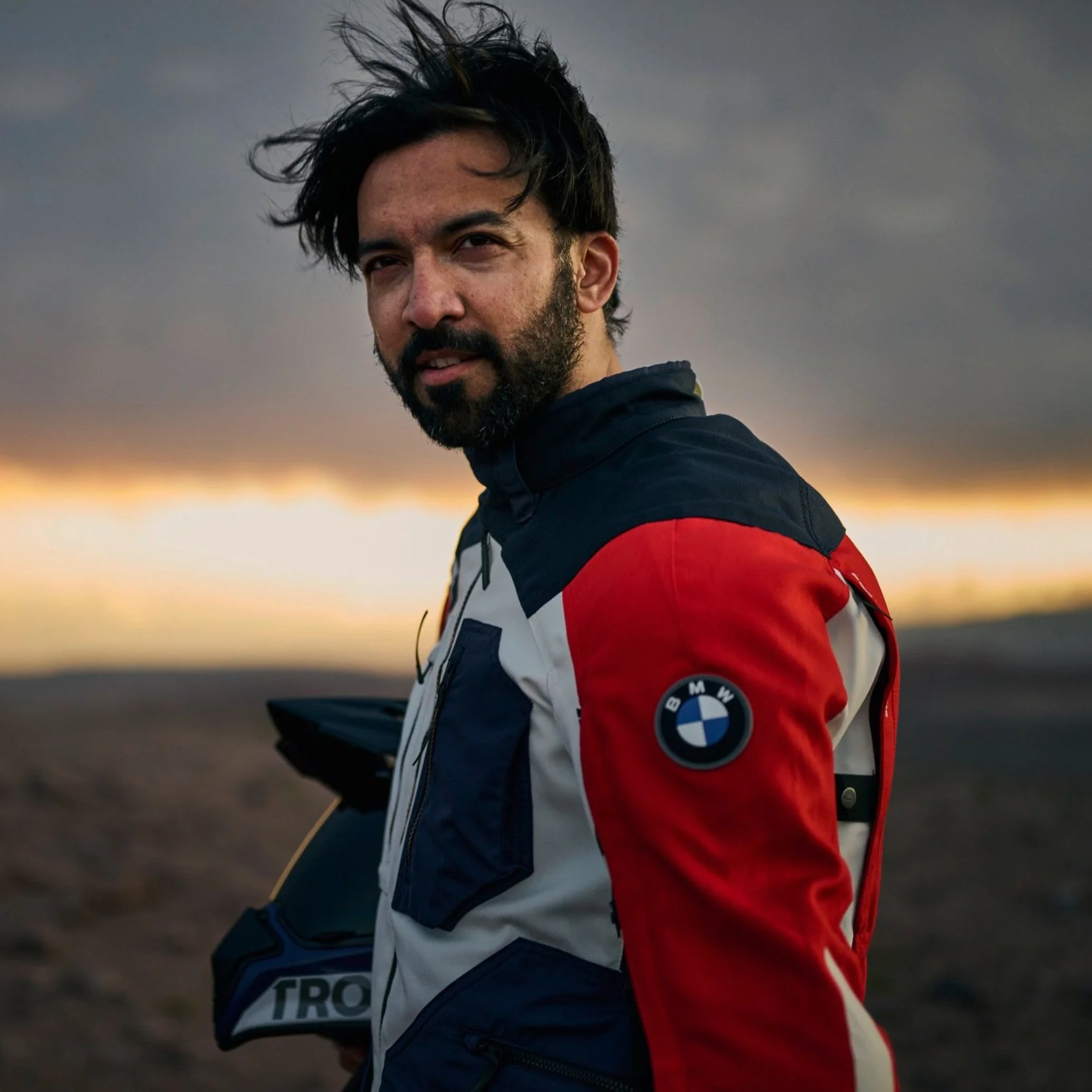 SYED SHAHNAWAZ KARIM wearing a BMW motorcycle jacket stands outdoors at sunset, holding a motorcycle helmet. The background features a cloudy sky with orange and yellow tones.