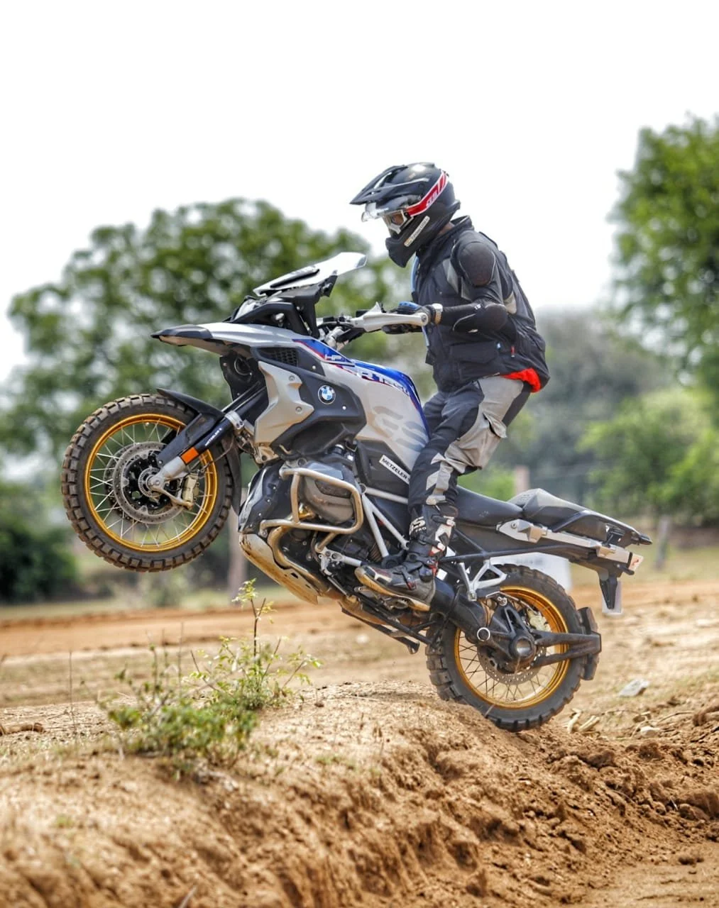 Syed SHAHNAWAZ KARIM performing a wheelie on an off-road adventure motorcycle with protective gear, on a dirt trail.