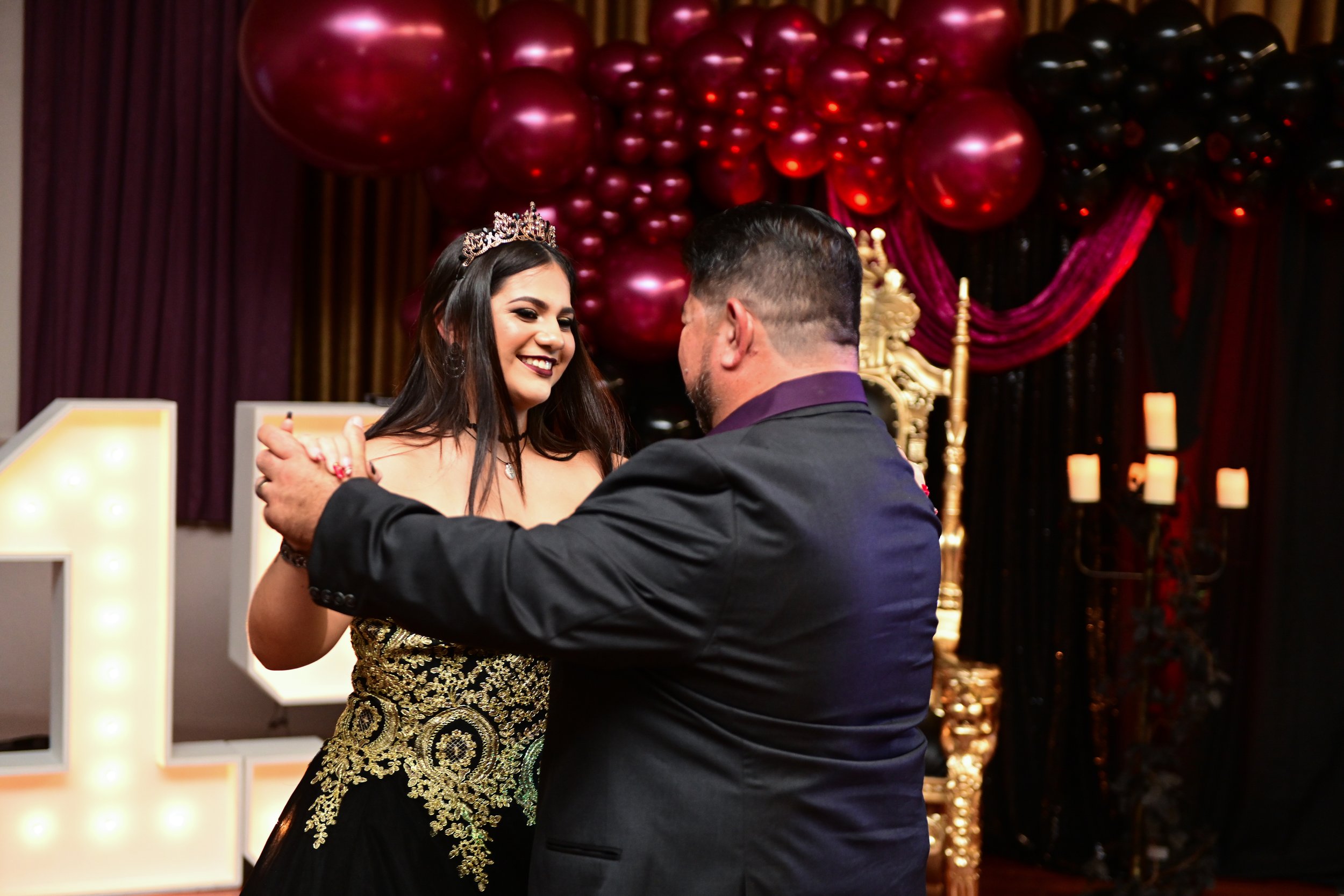 A woman wearing a tiara and a black and gold dress dancing with a man in a suit at a celebration event with balloons and candles in the background.