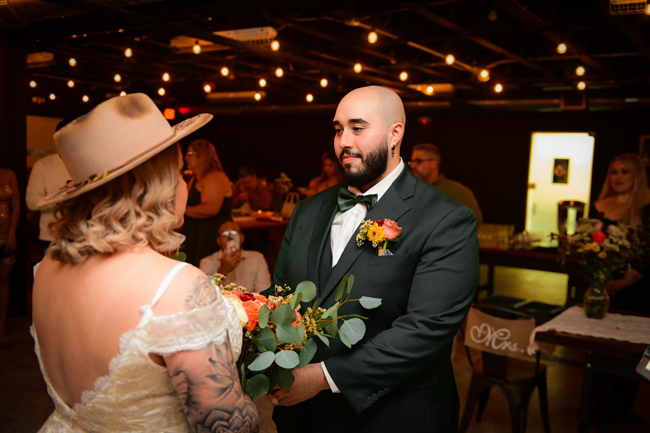 A wedding ceremony with a bald groom with a beard and cross earring in a black tuxedo, exchanging vows with a bride with long blonde hair, wearing a beige lace dress and wide-brimmed hat, indoors at a decorated venue with warm lighting, tables with f