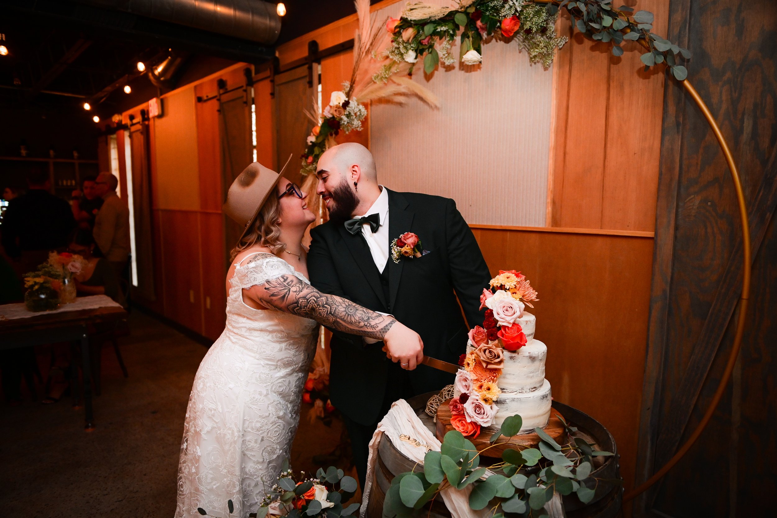 A couple eating cake at their wedding celebration, surrounded by floral and greenery arrangements in a warmly lit indoor venue.