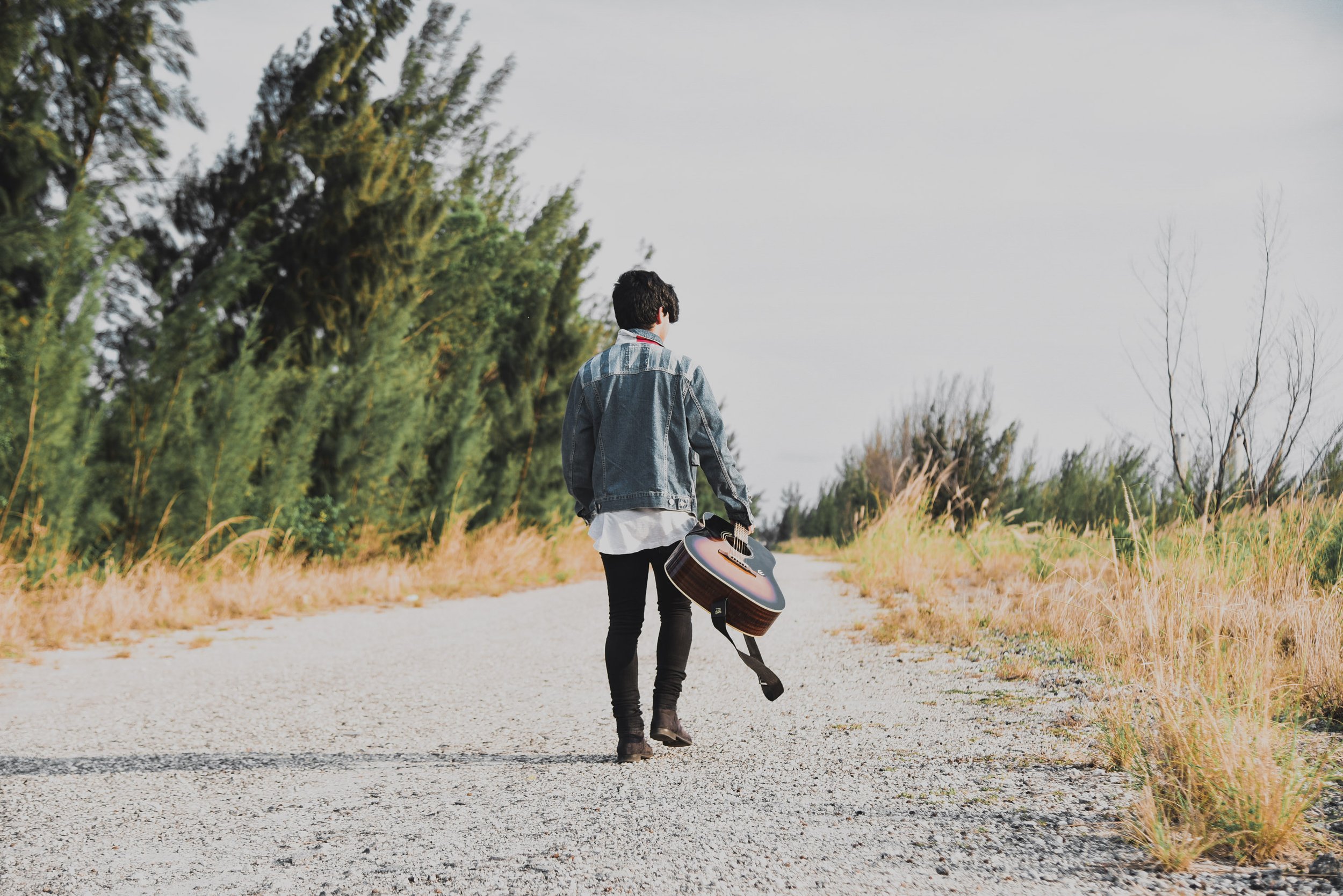 A person walking on a gravel path holding an acoustic guitar, surrounded by trees and grasses, with an overcast sky.