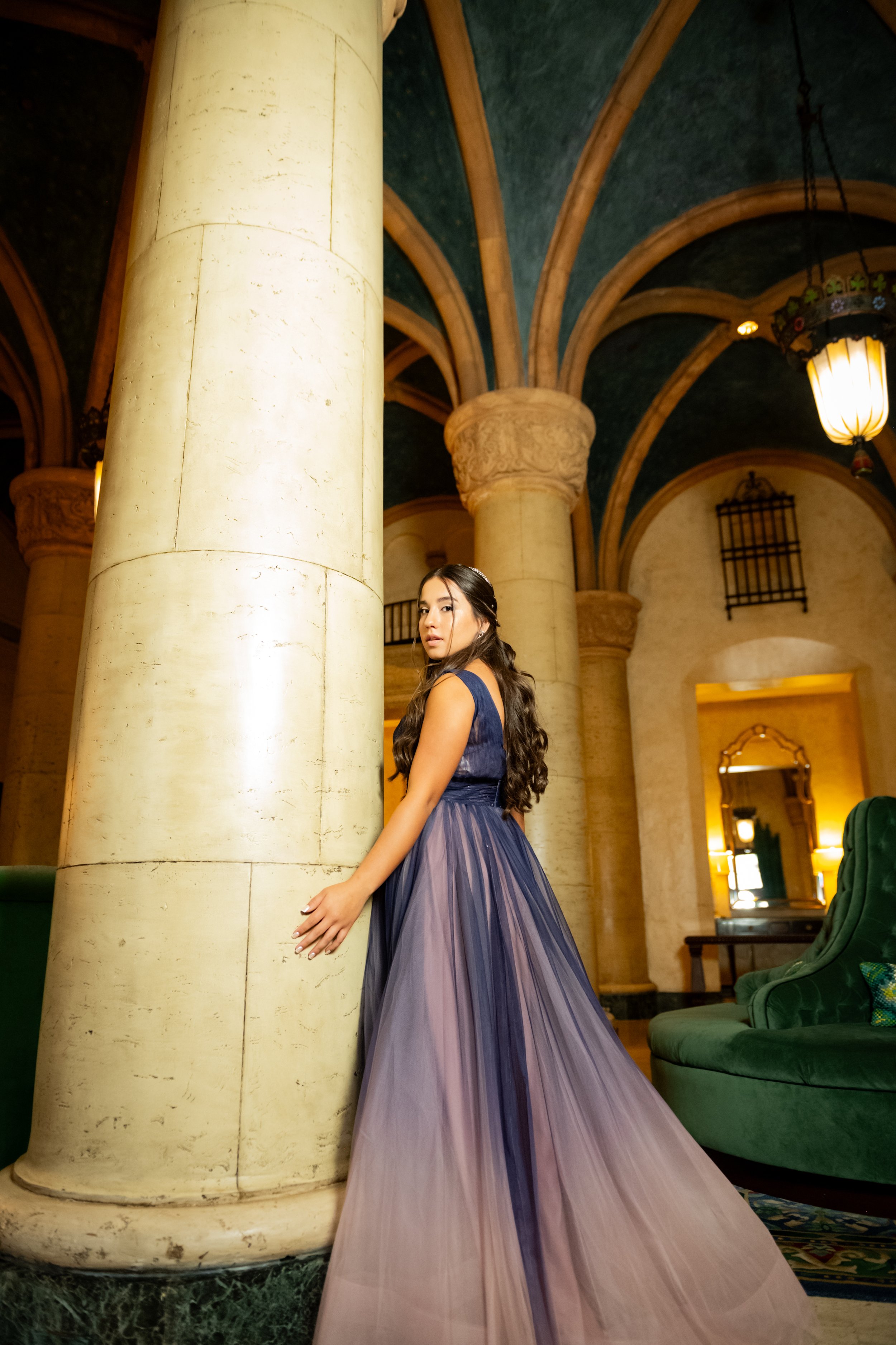 A young woman with long, dark hair, wearing a navy blue and purple evening gown, stands next to a large, beige marble column in an ornately decorated indoor space, possibly a hotel lobby or historic building.