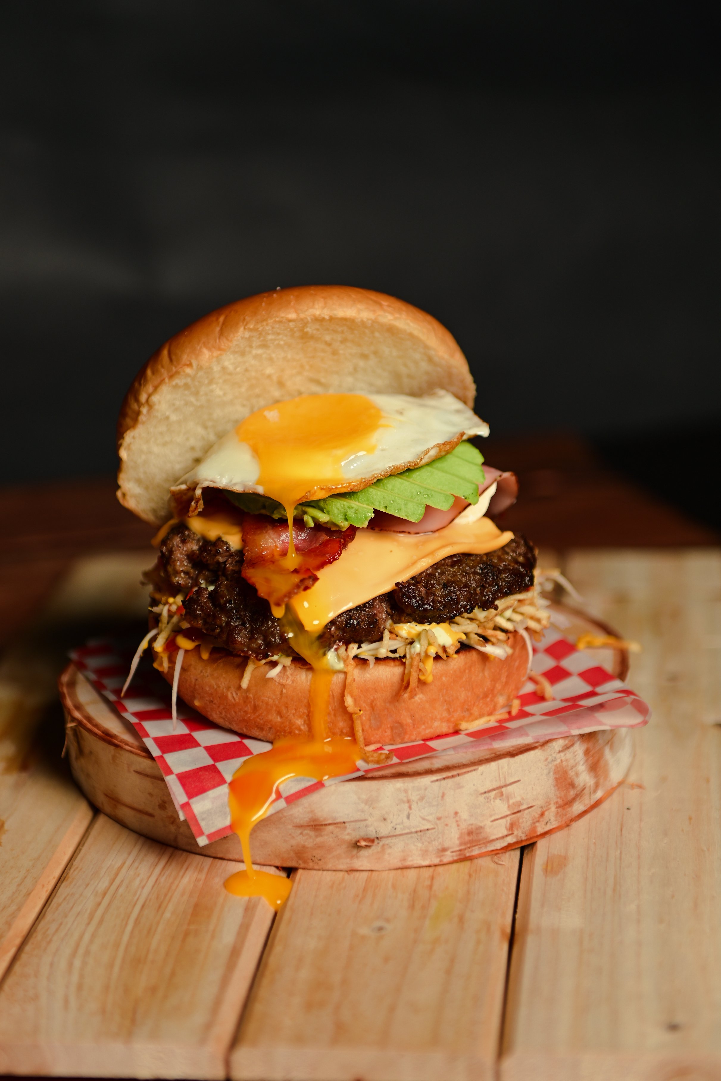 A cheeseburger with a fried egg, lettuce, tomato, bacon, and a beef patty on a toasted bun, with cheese dripping over the side, served on a checkered paper on a wooden surface.