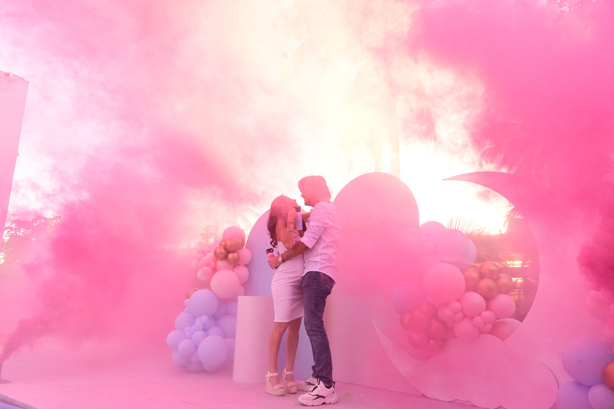 A couple sharing a kiss at an outdoor event with pink and white balloons and palm trees in the background during sunset.