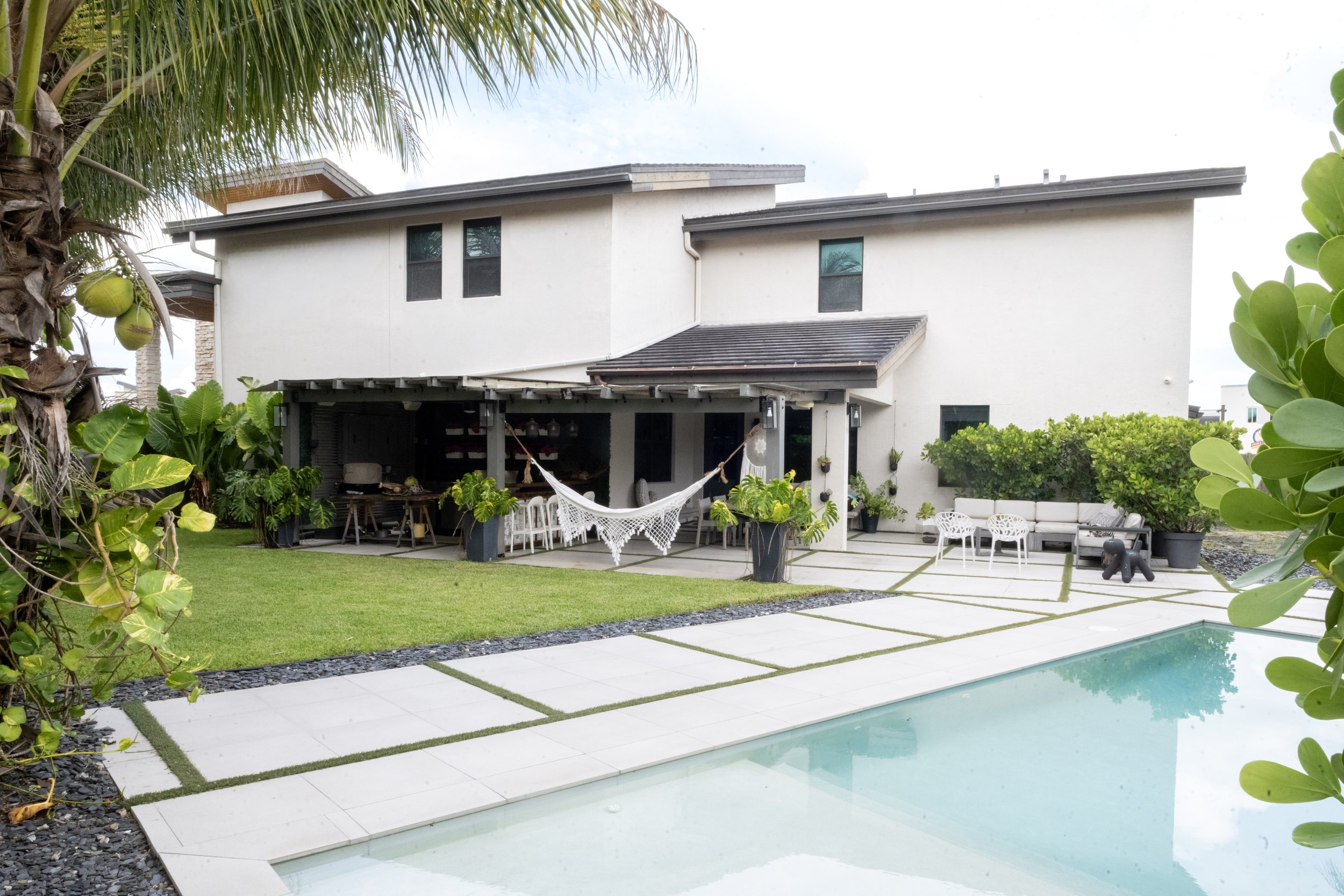 Backyard with a swimming pool, patio furniture, and lush greenery, including palm trees and bushes, against a white house.