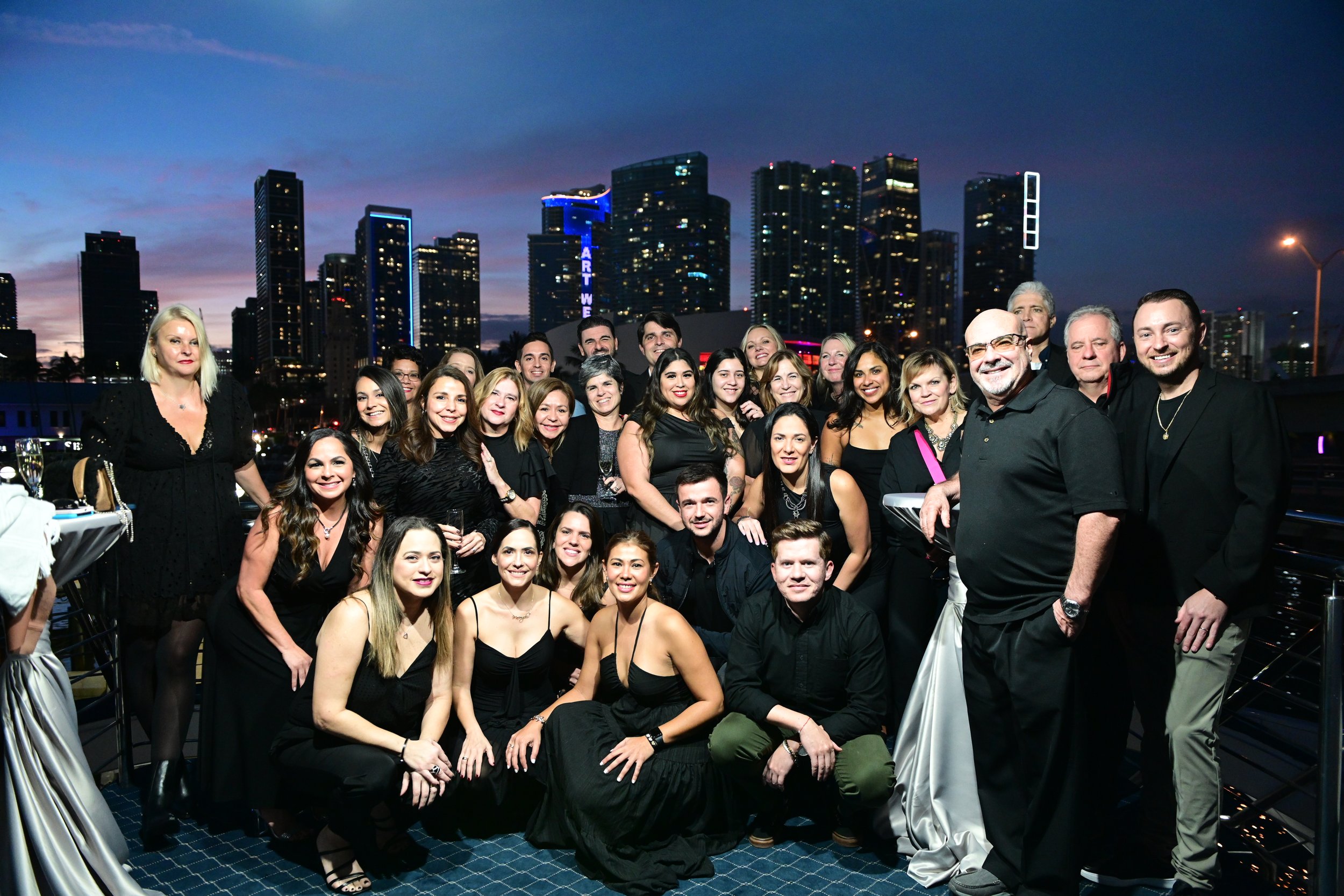 A large group of diverse people dressed in black attire celebrating outdoors during the evening with a city skyline and tall buildings illuminated in the background.