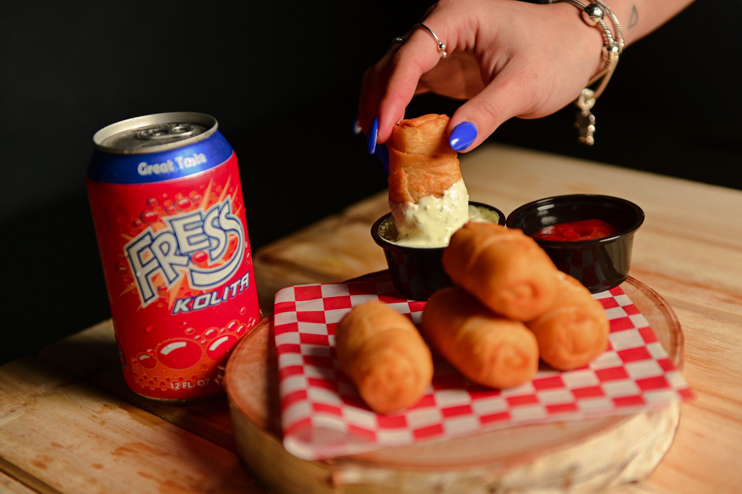 Close-up of a person's hand dipping mozzarella sticks into marinara sauce on a checkered paper-lined tray, with a can of Seltzer Kolita soda nearby.
