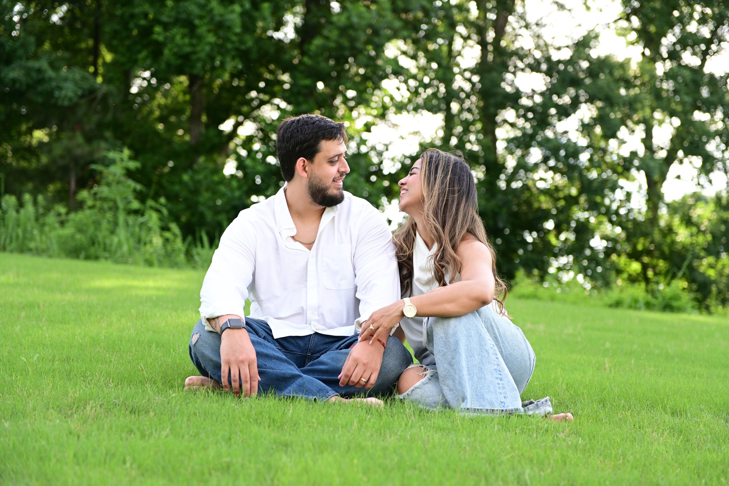 A man and a woman sitting on grass in a park, smiling and looking at each other with trees in the background.