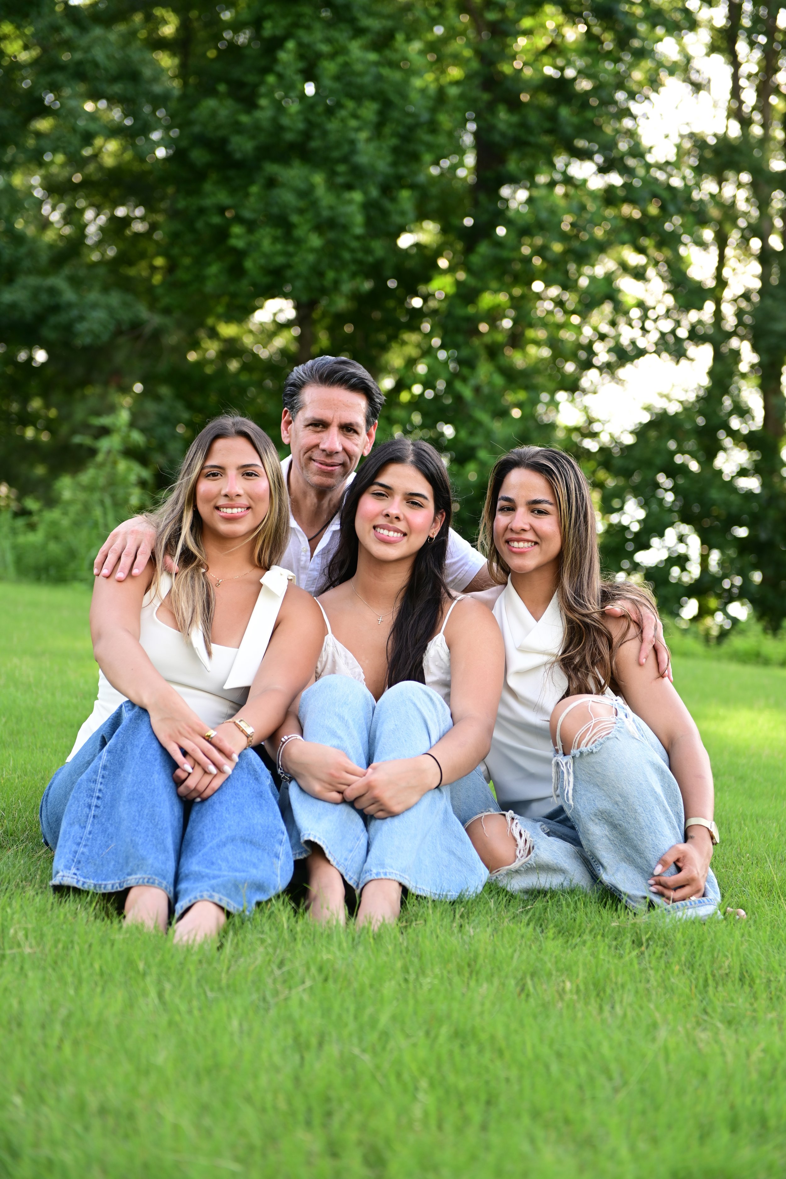 A man and three young women sitting on grass outdoors with trees in the background, smiling and posing for a photo.