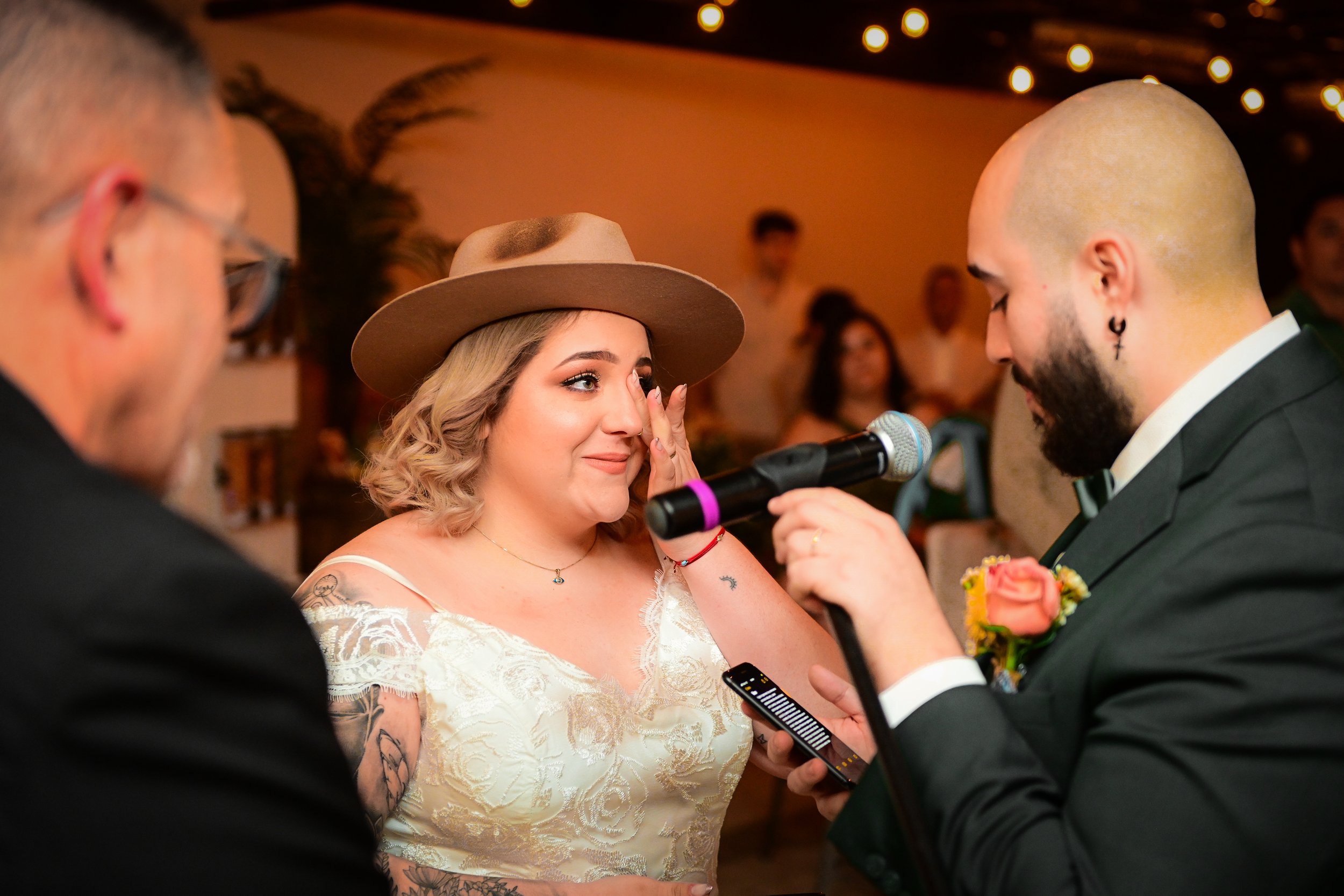 A couple is getting married, with the bride wiping away tears and the groom holding a microphone for the bride to speak. The bride is wearing a white lace wedding dress and tan hat, and the groom is in a black tuxedo with a boutonnière. The backgroun