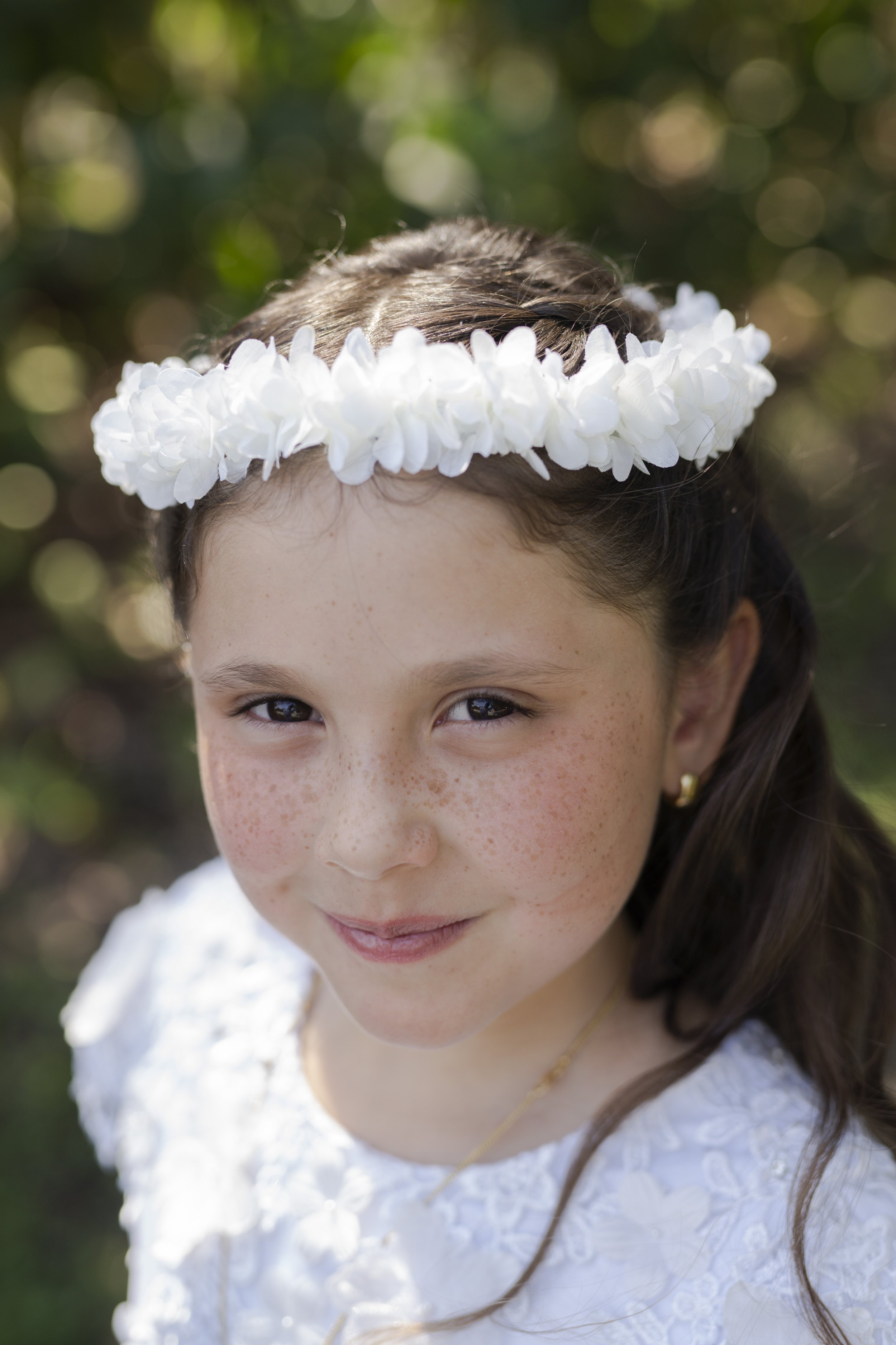 Close-up of a young girl with brown hair, freckles, and brown eyes, wearing a white floral headband and white dress, outdoors with blurred greenery in the background.